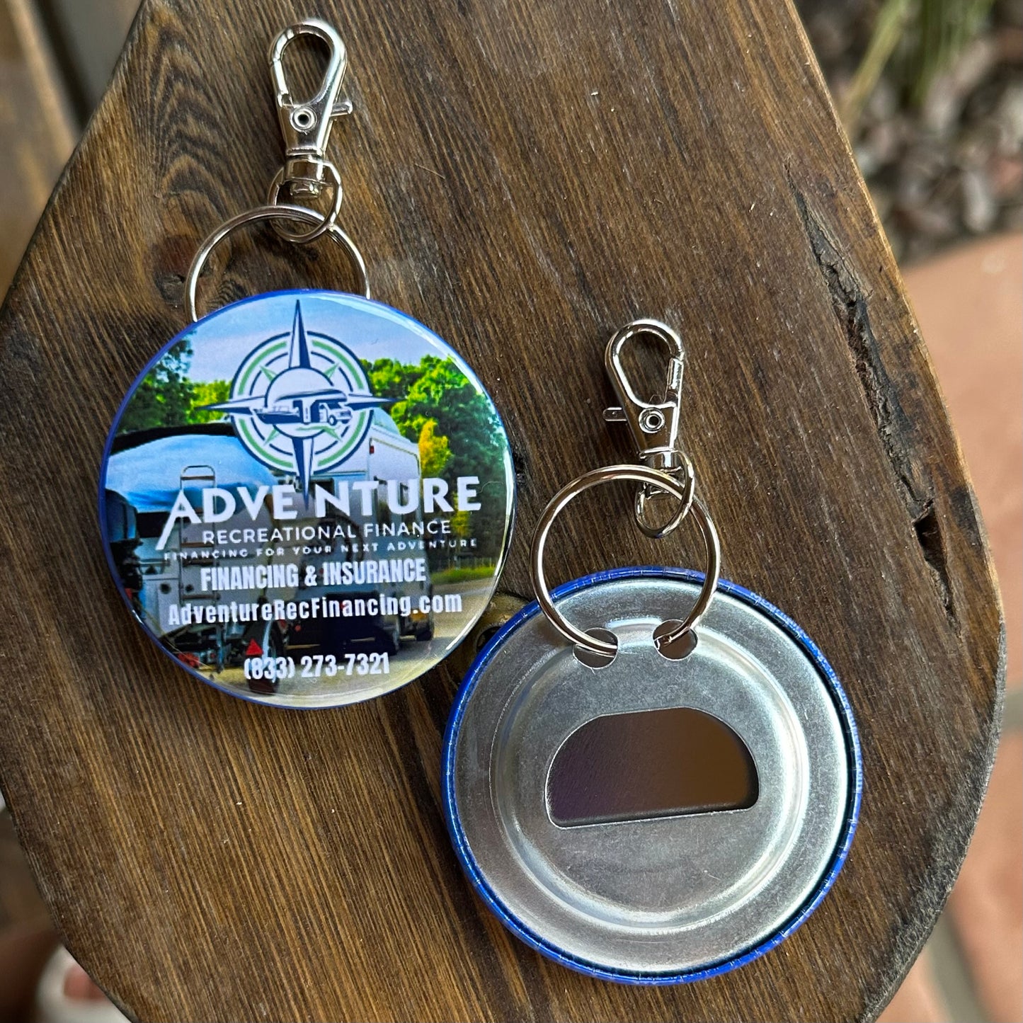 Two keychain items on a wooden surface, one with the reverse side of a bottle cap opener and the other with a logo and text.